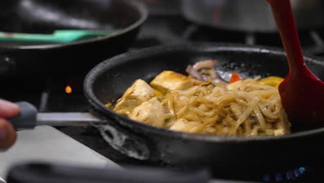 la espátula agita el plato de tofu de fideos de arroz plano chisporroteante sobre la llama caliente en la cocina del restaurante comercial, el chef sacude y lanza la almohadilla vegetariana kee mao, cámara lenta de primer plano 4k