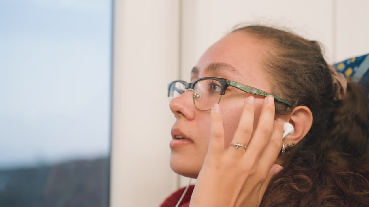 Close up young woman wearing glasses listening to earbuds by window, relaxed gaze drifts toward passing landscape, subtle smile and earbud adjustment convey content evening commute mood