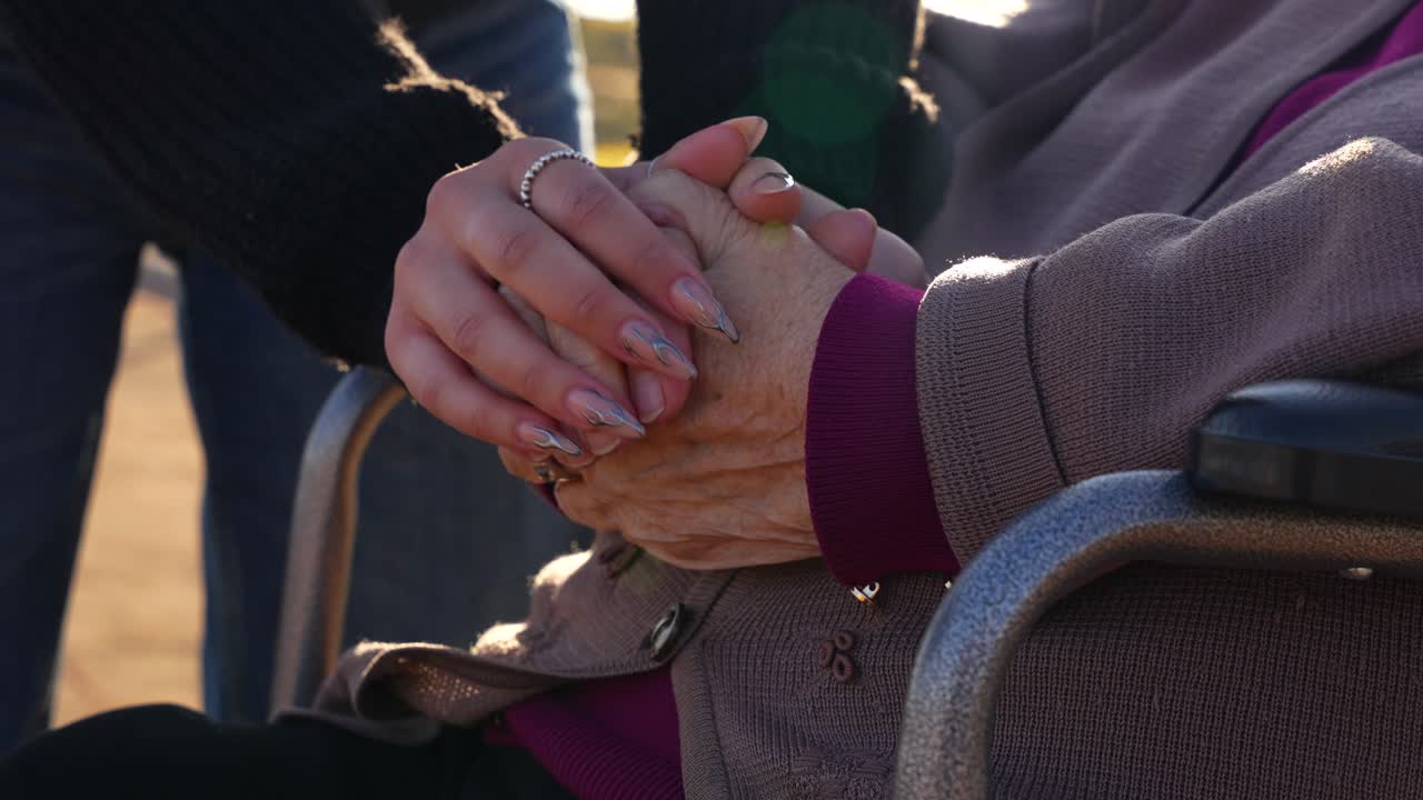 Granddaughter holding the wrinkled hands of her wheelchair-bound grandmother to warm her up. Close up