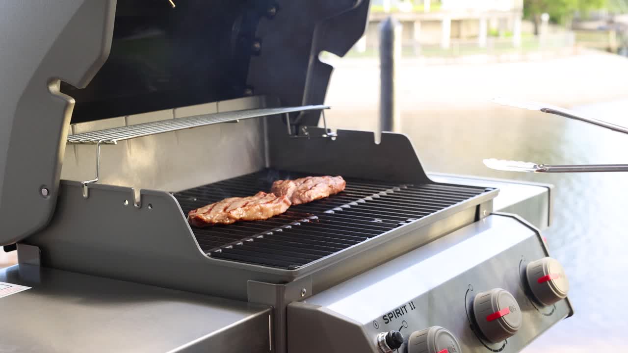 A person grills beef steaks on a barbecue by the water, showcasing outdoor cooking techniques and a serene environment