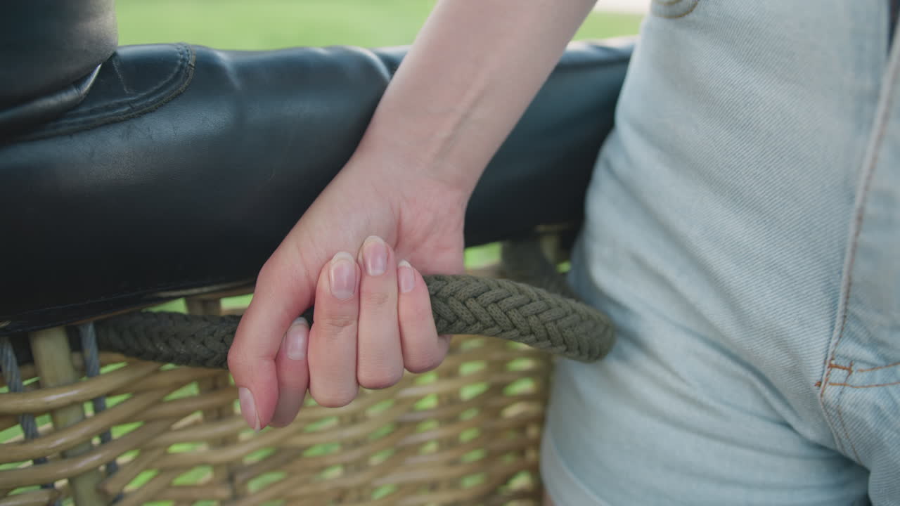 Close up of hand tightly gripping braided rope inside woven wicker basket, denim fabric visible beside black leather cushion, suggesting tension or support during balloon ride under soft daylight