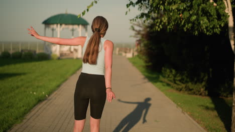 back view of young lady skating outdoors, arms wide open for balance on sunny day, surrounded by lush greenery, trees, and blurred view of people walking in background with gazebo visible in distance