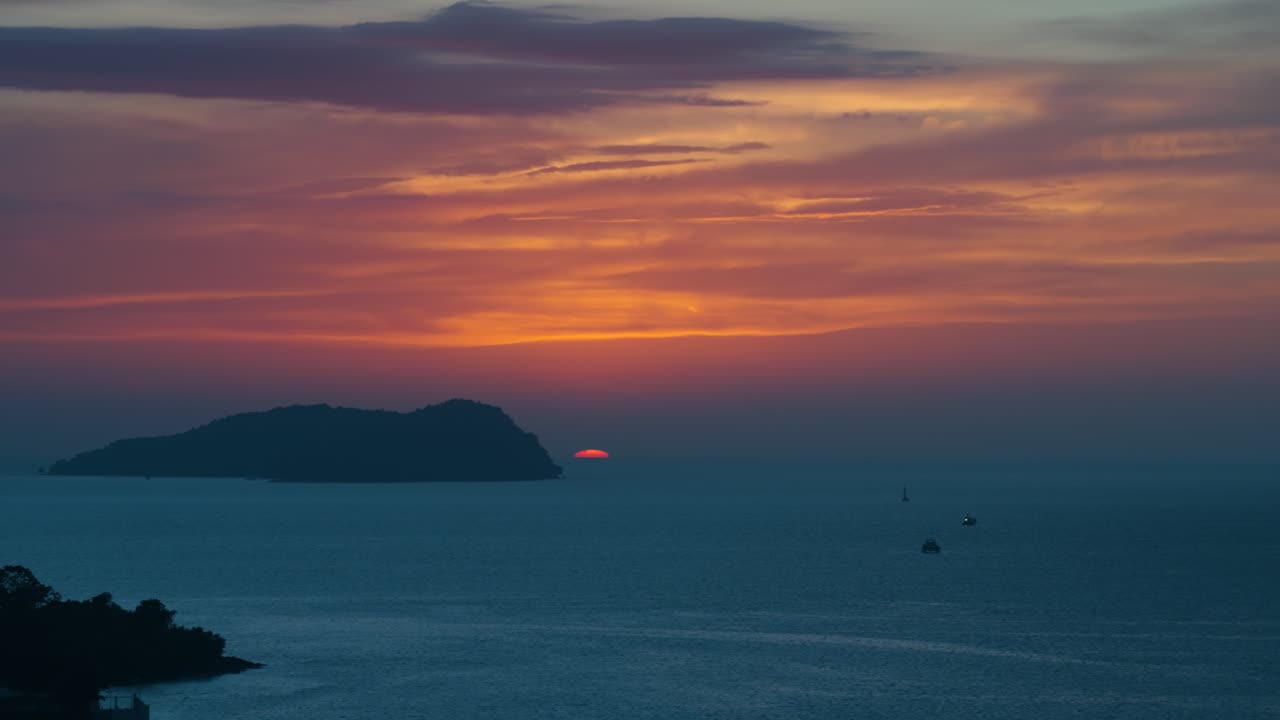 Aerial close-up captures the red sun setting behind a silhouetted island and seascape in Kota Kinabalu under a vibrant orange and blue sky as a bird flies by