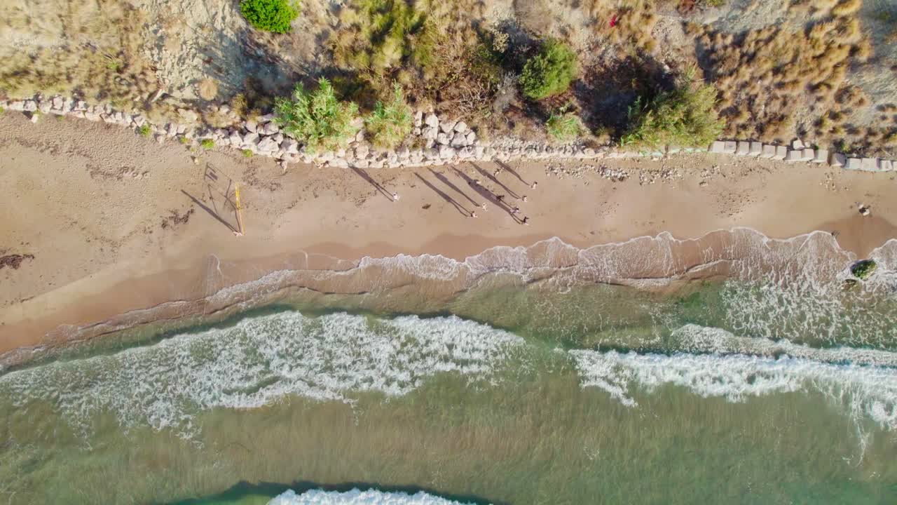 Crashing Waves Onto Sandy Shores With Tourists Strolling On A Sunny Day. Aerial Topdown Shot