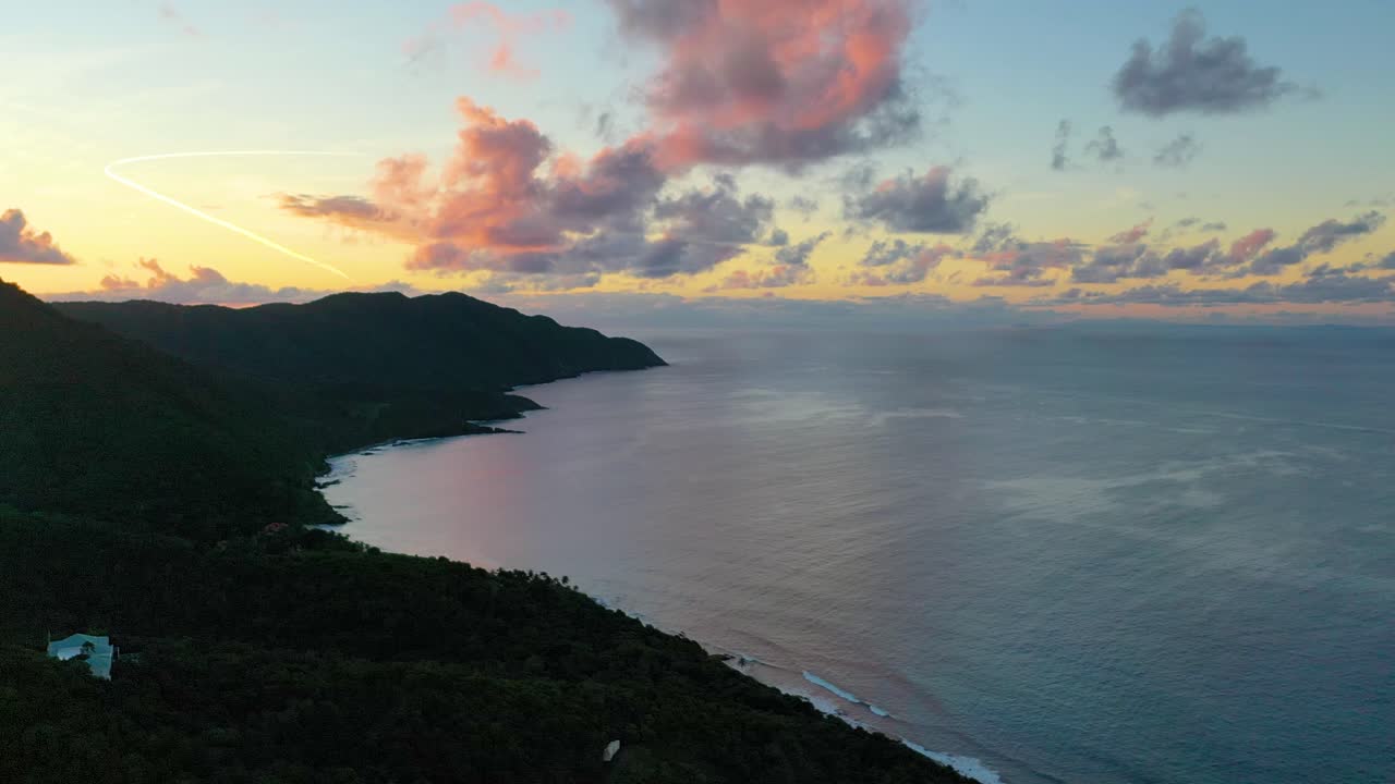 Drone footage captures the tranquil Caribbean Sea near St. Croix at dawn, with a rich, colorful sky illuminating a tree-covered peninsula and casting soft reflections on the shore