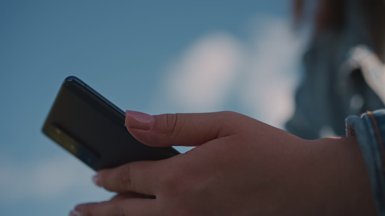 Persona che usa un dispositivo mobile alla luce del sole. Un individuo che naviga sullo smartphone sotto un cielo sereno e una giornata luminosa. Una persona interagisce con lo smartphone all'aperto sotto un cielo aperto con nuvole e sole.