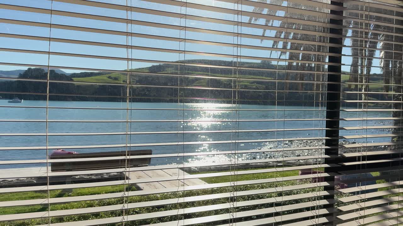 A tranquil lakeside scene viewed through window blinds, capturing sunlight reflections on water in Akaroa, New Zealand