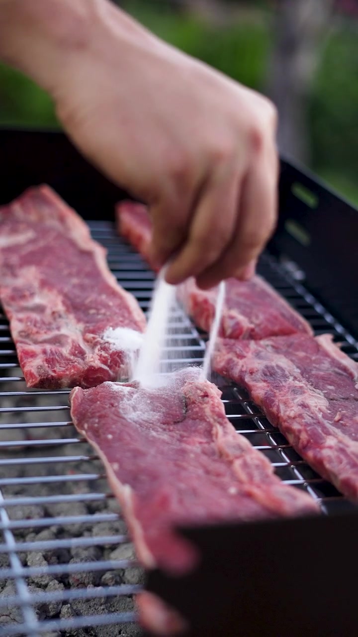 Vertical video of hand sprinkling salt on raw beef ribs on grill