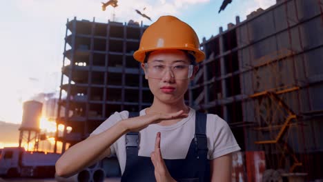 Close Up Of Asian Woman Worker Wearing Goggles And Safety Helmet Looking At Camera And Showing Time Out Hands Gesture While Standing At Construction Site