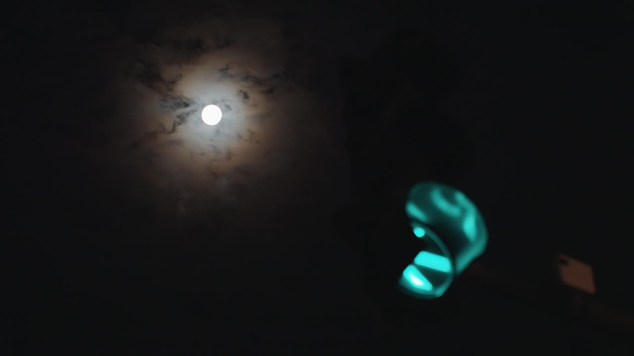 Luna llena sobre semáforo verde, halo a través de nubes finas, poste en silueta e intersección urbana, bucle cinemático lento que sugiere entrega nocturna y trayecto vacío, atmósfera luminosa y melancólica.