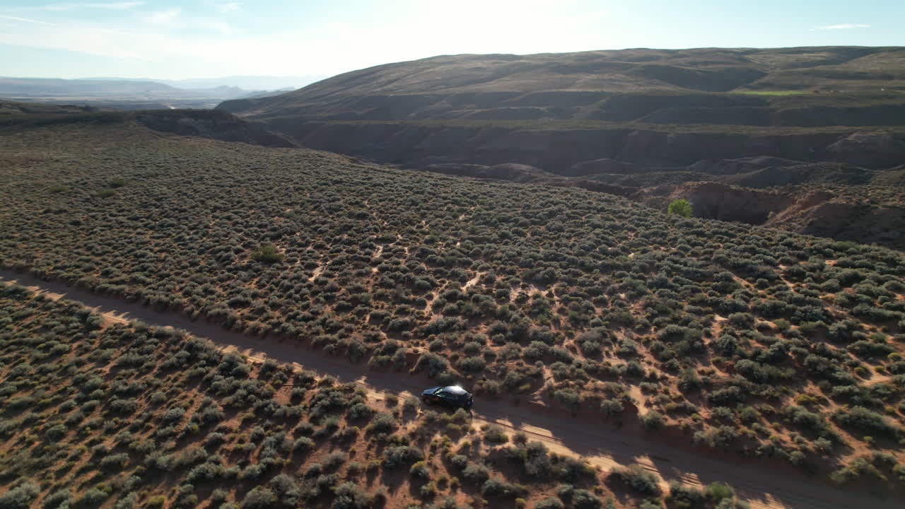 SUV traveling along a dirt road through a rugged Utah desert landscape, surrounded by bushes and hills in the distance.