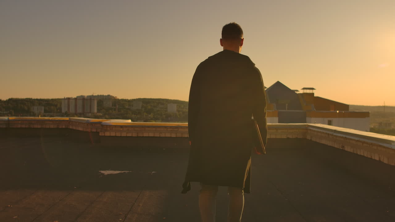 The camera follows a man walking across a rooftop at sunset with a laptop and a bottle. The freelancer opens the laptop and types on the keyboard. Slow motion hacker walks on the roof with a laptop.