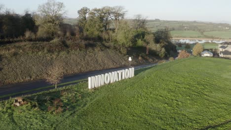 Large-white-innovation-sign-in-roadside-green-pasture,-sunny-aerial