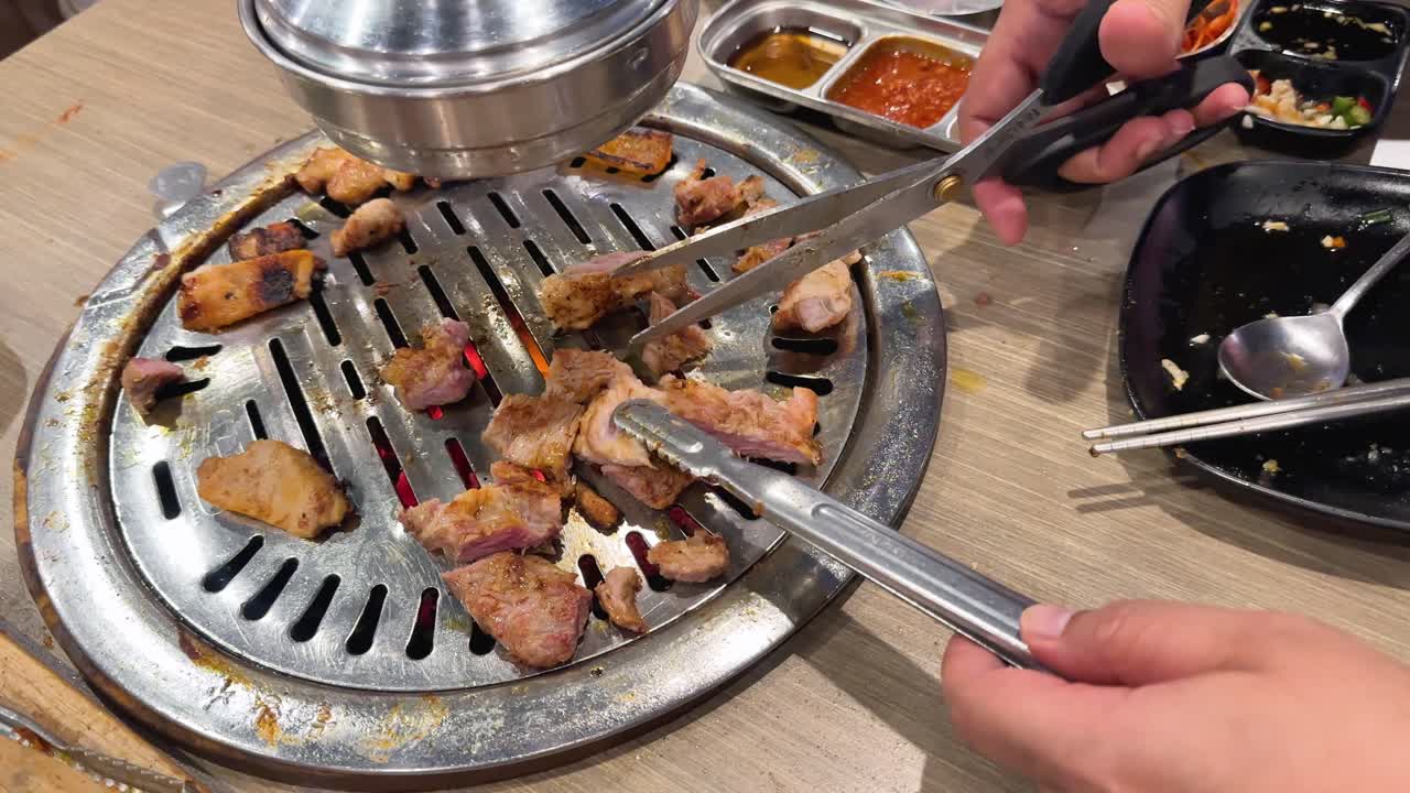 Hands using scissors and tongs to cut grilled pork on a BBQ grill. Bright lighting, close-up view, lively dining atmosphere