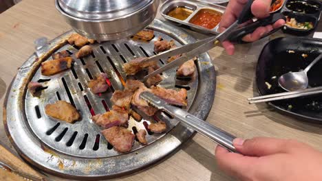 hands using scissors and tongs to cut grilled pork on a bbq grill. bright lighting, close-up view, lively dining atmosphere