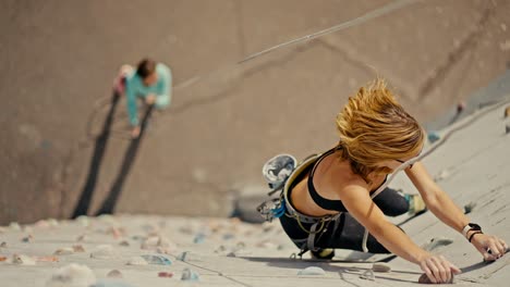 una chica rubia en un uniforme de verano deportivo negro sube a una pared de escalada blanca con repisas multicolores y es asegurada por su amiga en una chaqueta azul que está de pie en el suelo debajo y sostiene una cuerda. vista superior de una chica rubius subiendo a una muralla de escalada y practicando escalada en roca