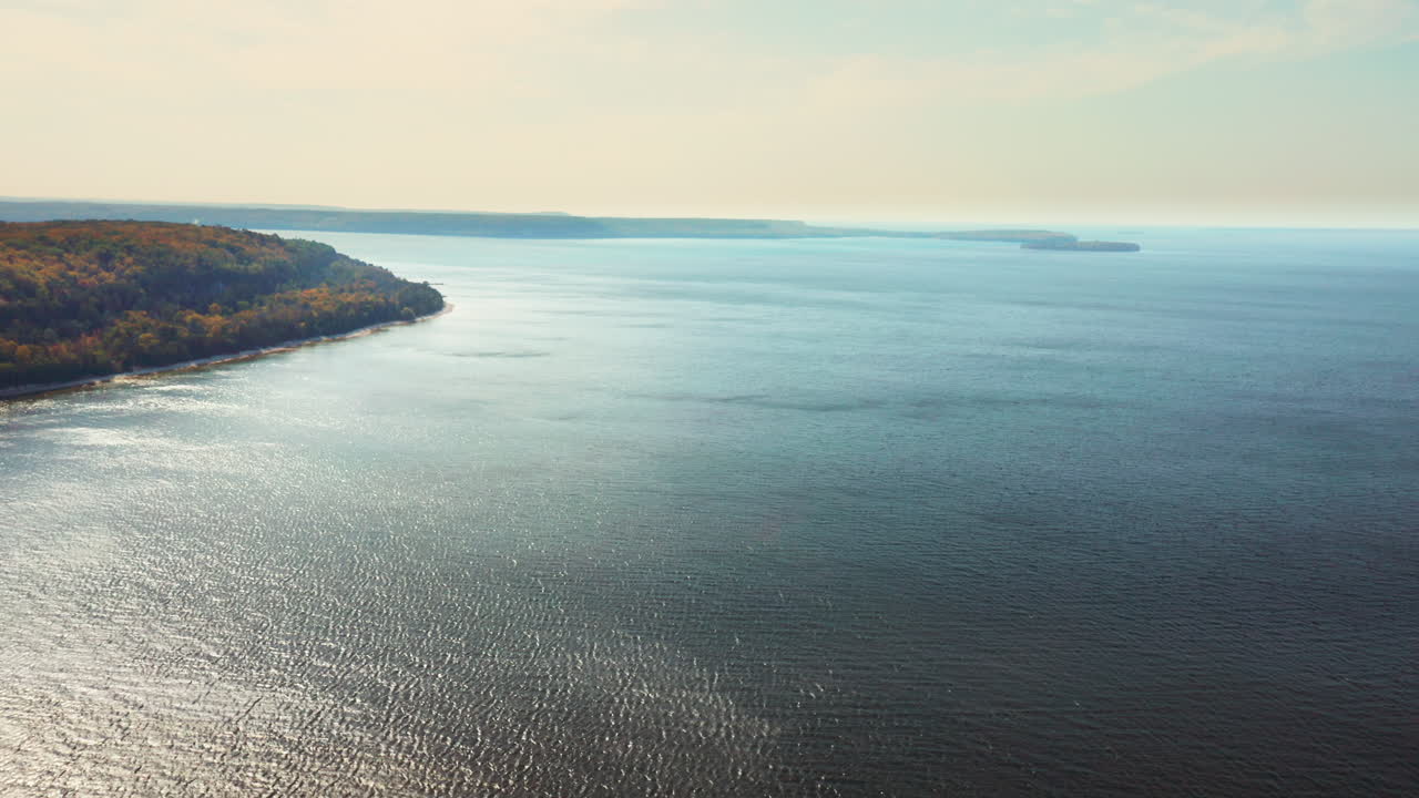 Gentle waves shimmer beneath a pale sky as autumn-colored forests trace the lake’s curved edge, where sunlight glows across the open water in calm serenity
