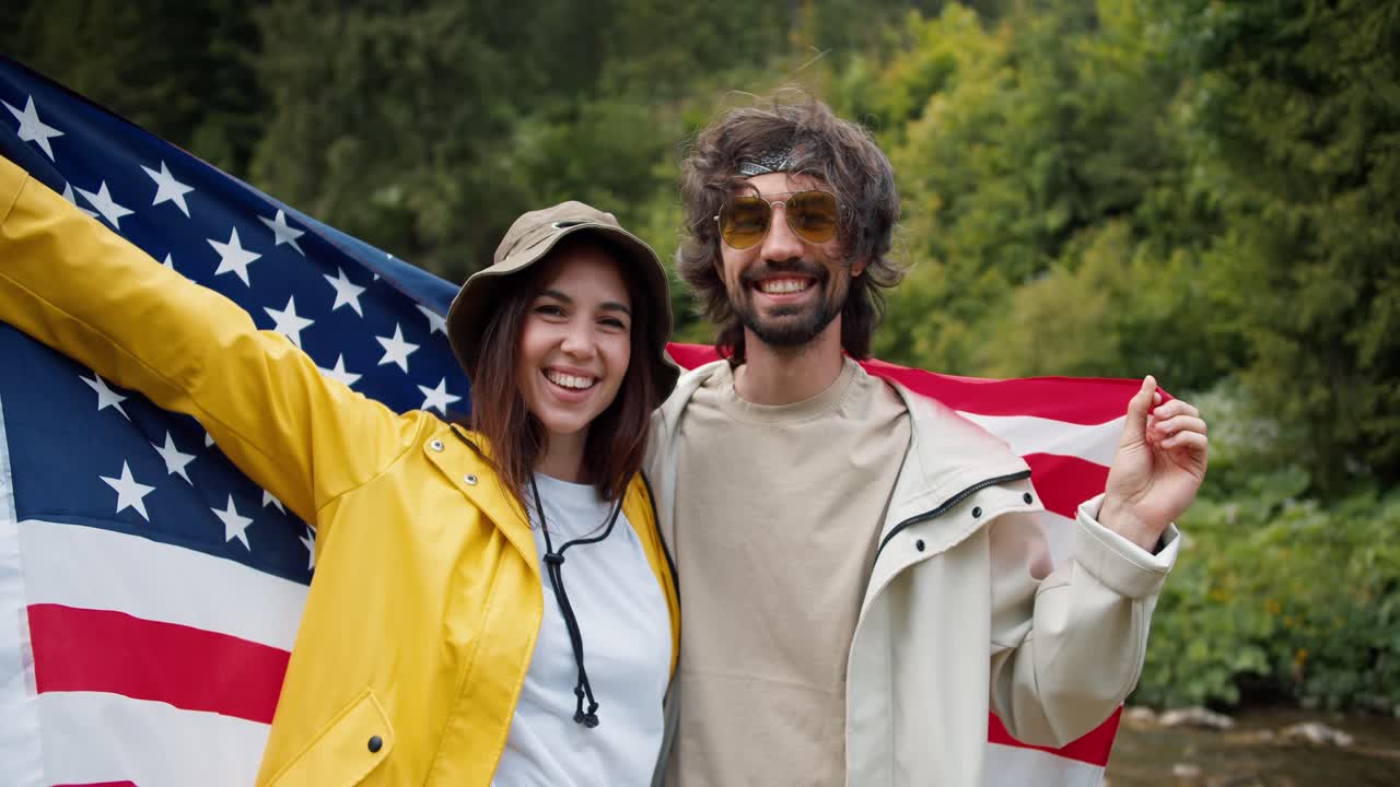 The brunette guy and girl pose and admire, they raised the US flag during their trip through the mountain forests against the background of green trees
