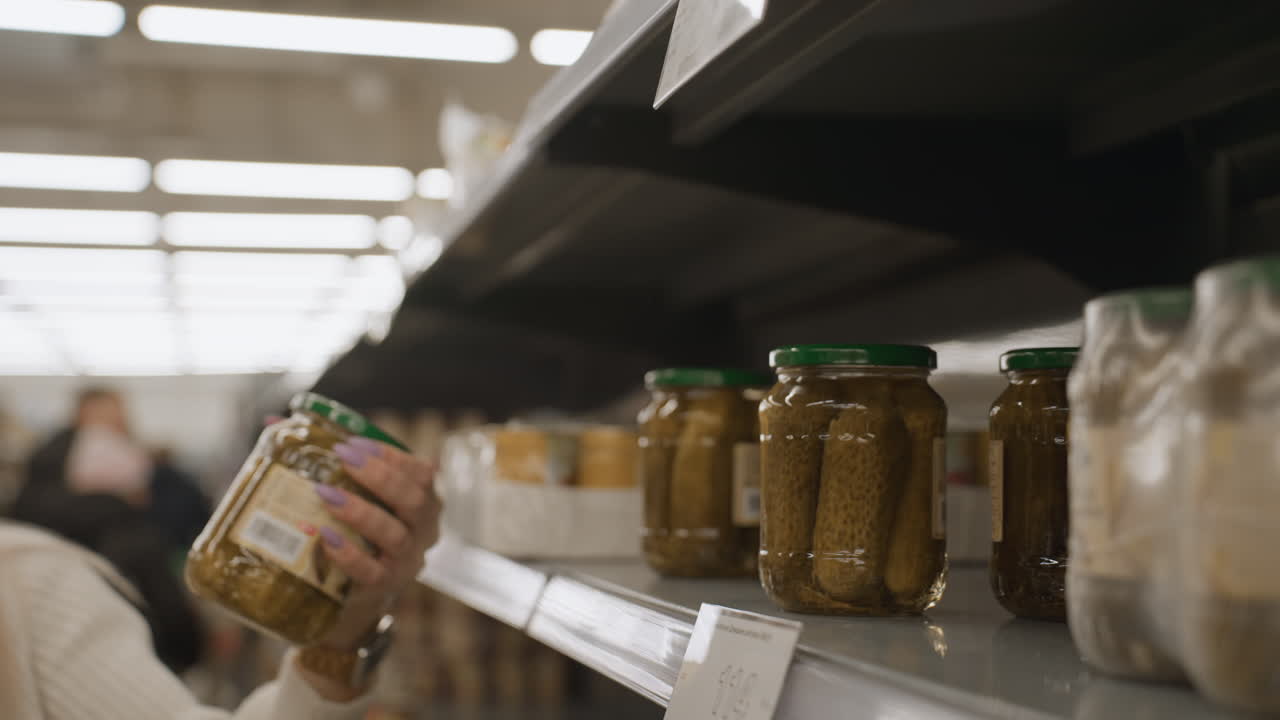 Close up view of shopper hand reaching for glass jar of pickled cucumbers on supermarket shelf in well lit aisle showcasing jar label design during grocery selection with blurred product background