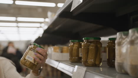 close up view of shopper hand reaching for glass jar of pickled cucumbers on supermarket shelf in well lit aisle showcasing jar label design during grocery selection with blurred product background