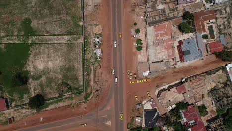 Vista-Aérea-De-Pájaro-Siguiendo-Una-Camioneta-Blanca-En-La-Carretera-Kololi-En-Serrekunda-África-Con-Caminos-Polvorientos-Rojos