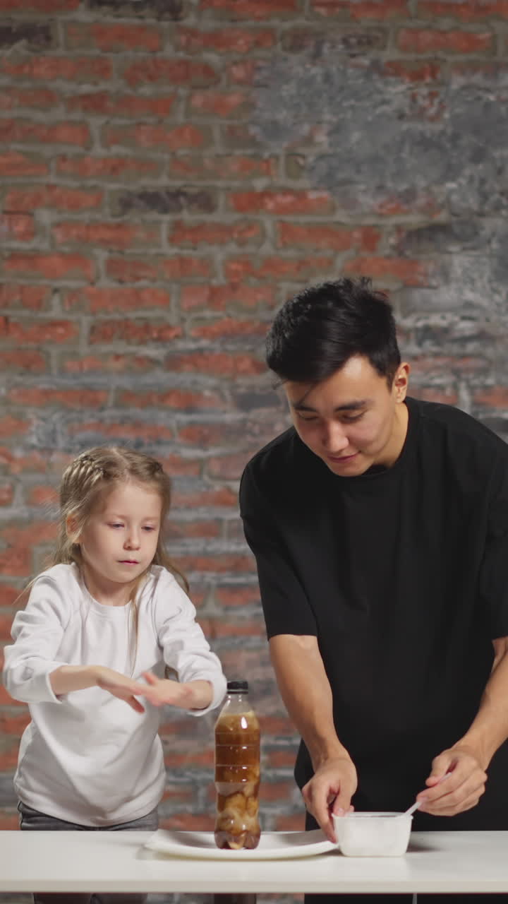 Shocked little girl tries to stop splashing soda drink and jumps away from young Asian teacher conducting scientific experiment in home lab slow motion