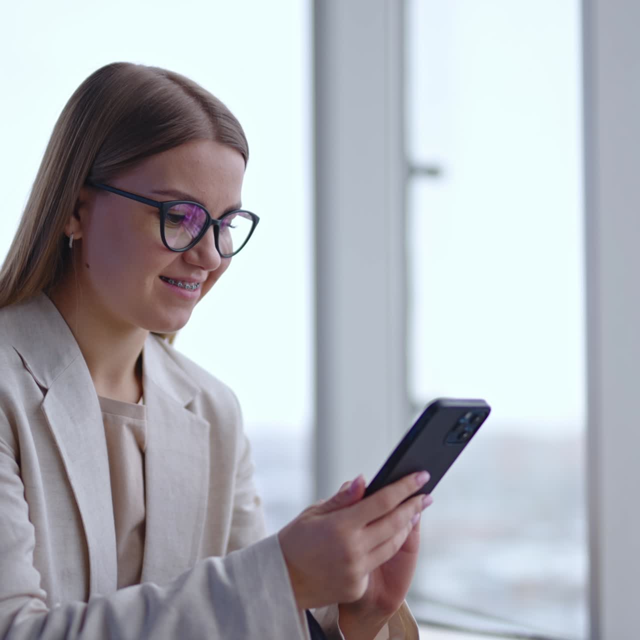 Beautiful young lady in glasses searches information on the phone. Woman looks at laptop and then opens her notebook. Light panoramic windows backdrop
