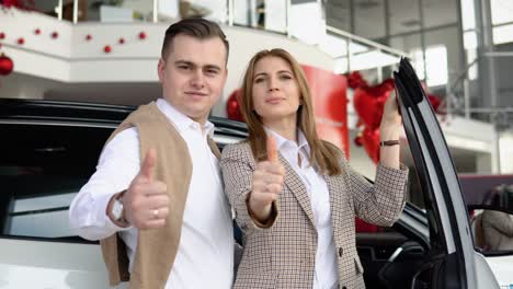 happy couple at a car dealership with a new car in the background. the guy gave his girlfriend a car for valentine's day