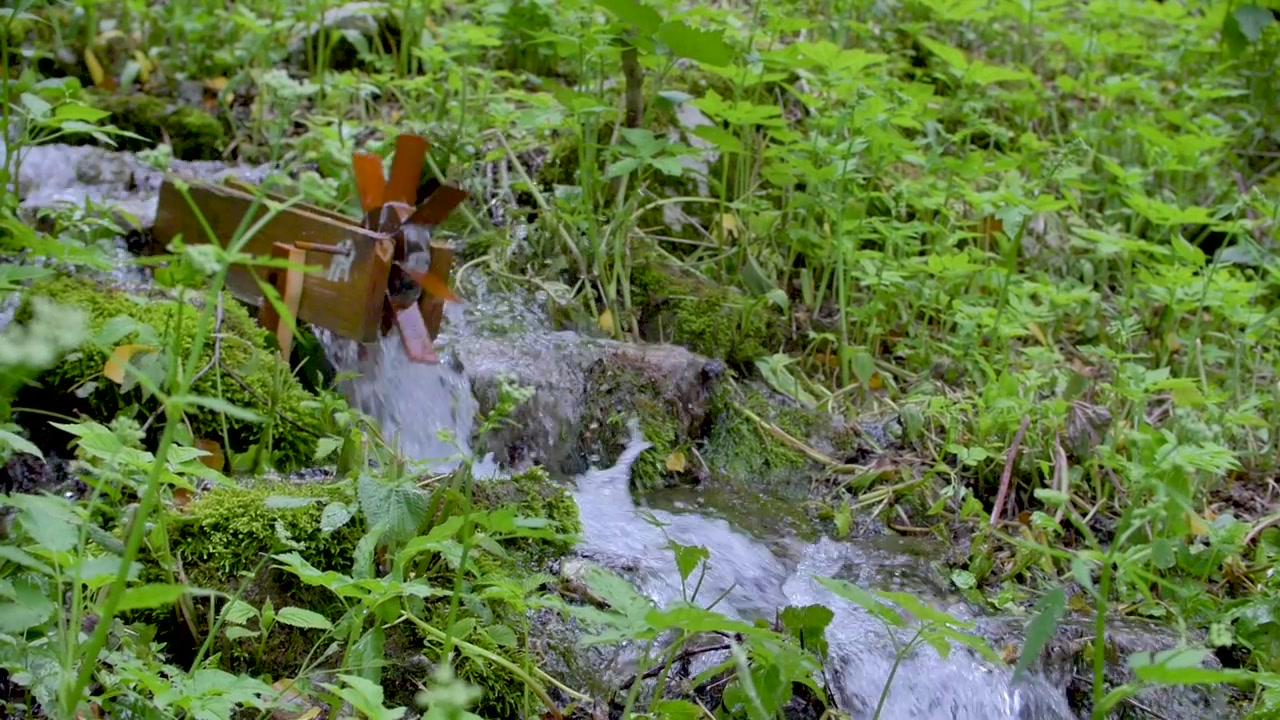 Small wooden water mill rotating on small creek down the hill in the green undergrowth.Water wheel in top left corner of frame.