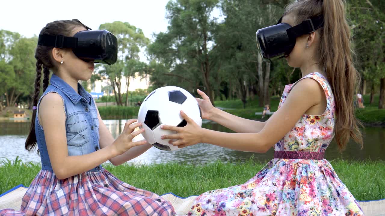 dos niños juegan juegos virtuales. están sentados en gafas de realidad virtual en el césped de la orilla del río, con una pelota de fútbol en las manos y girándola.