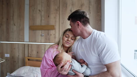 happy couple sit on a hospital bed holding their newborn baby. new-made parents with infant child.