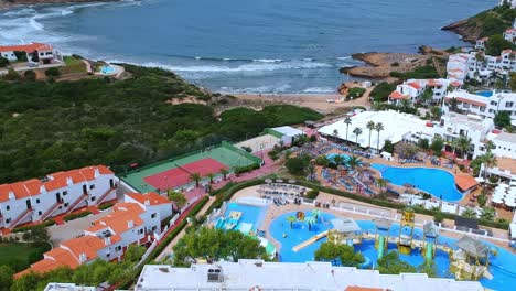 Vistas-Aéreas-De-Gente-De-Vacaciones,-Jugando-Tenis,-En-La-Piscina-Y-Relajándose-En-La-Playa-En-Menorca,-España.