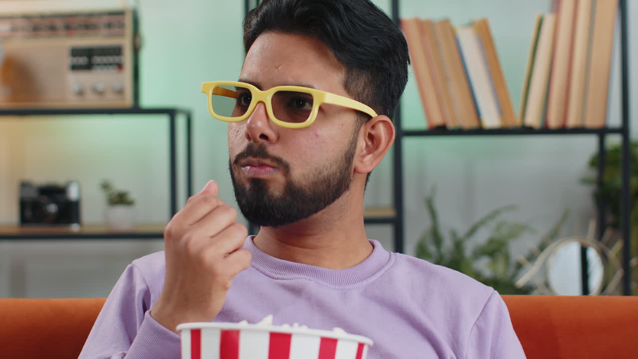 Indian man sits on couch eating popcorn and watching interesting tv serial sport game online at home