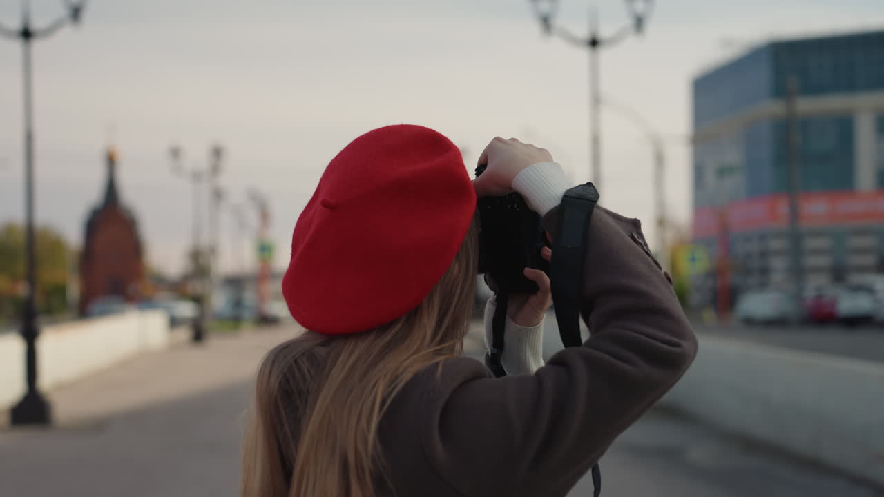 Giovane donna bianca con basco rosso che cammina lungo un ponte cittadino con un cappotto vintage e una macchina fotografica a mano, guglia di chiesa in lontananza, cielo coperto, passo contemplativo, morbide tonalità autunnali, ritratto di viaggio spontaneo che cattura