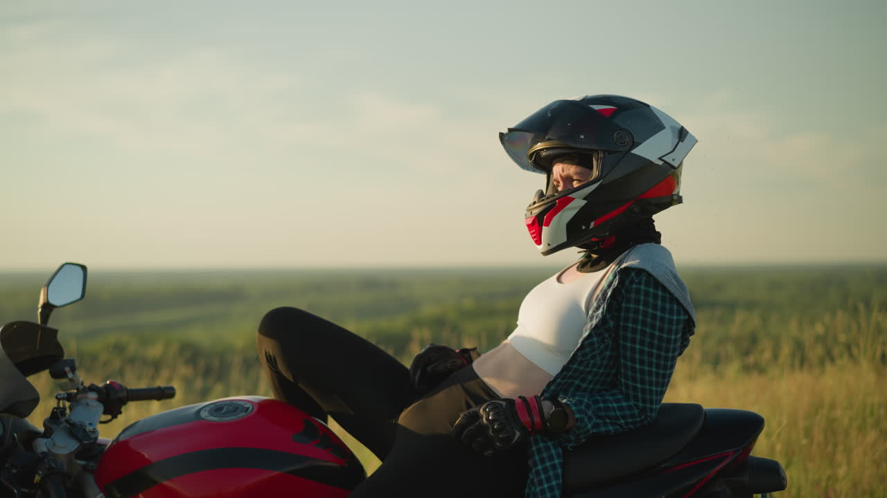 una mujer joven con casco descansa en una bicicleta eléctrica roja en un campo abierto, mirando fijamente a algo, su tapa blanca es visible mientras se inclina hacia atrás en la motocicleta