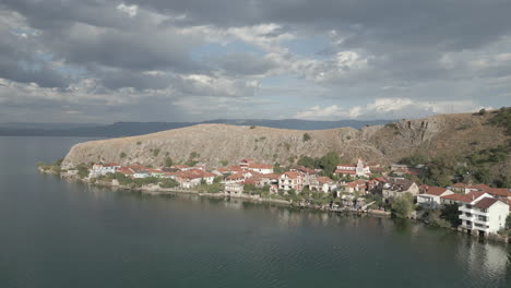 Droneshot-from-above-Lin-Albania-flying-backwards-on-a-cloudy-day-with-the-sun-coming-through-the-clouds-with-land-and-water-underneath-LOG