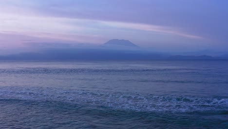 Vista-Aérea-De-Las-Olas-Que-Vienen-Hacia-La-Cámara-Y-El-Volcán-Agung-En-Segundo-Plano-Al-Atardecer