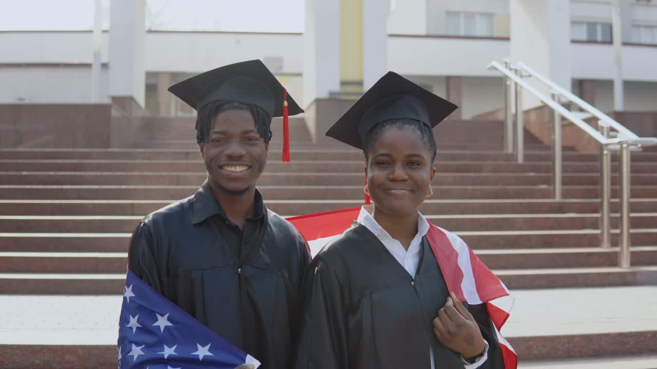 un hombre y una mujer afroamericanos se paran uno al lado del otro frente a la cámara con túnicas negras y sombreros cuadrados de estudiantes graduados con la bandera de los estados unidos en los hombros