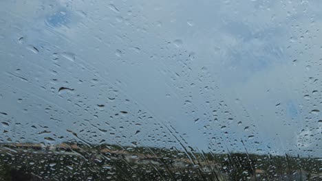 Nahaufnahme-Von-Regentropfen,-Die-Auf-Windschutzscheibe-Und-Fenster-Mit-Häusern-Und-Wolken-Im-Hintergrund-Fallen