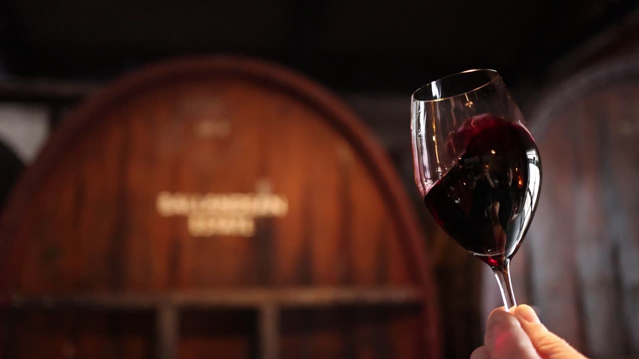Mano che fa roteare un bicchiere di vino rosso davanti a una botte di rovere nell'illuminazione calda di una cantina, al rallentatore