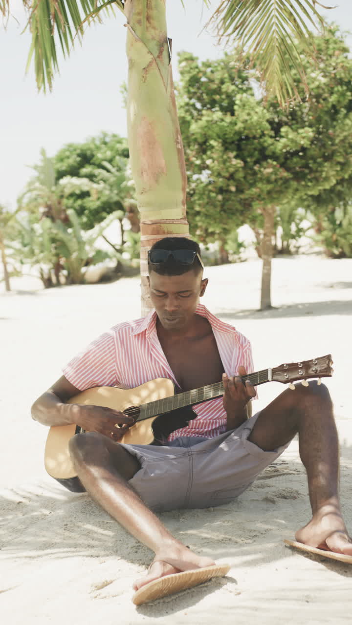 Vertical video of happy african american man playing guitar and singing on sunny beach, slow motion