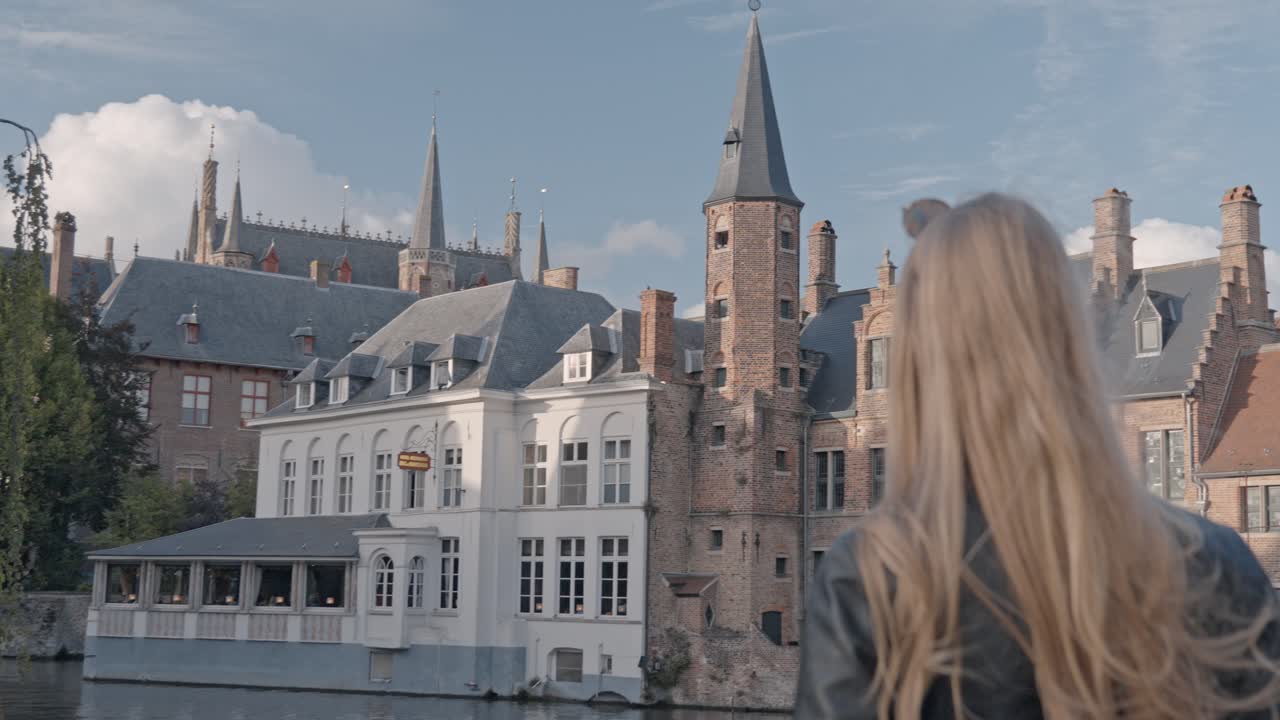 A beautiful blonde woman stands with her back to the viewer, gazing out over the tranquil canal at the iconic Rozenhoedkaai (Rosary Quay) in Bruges, Belgium