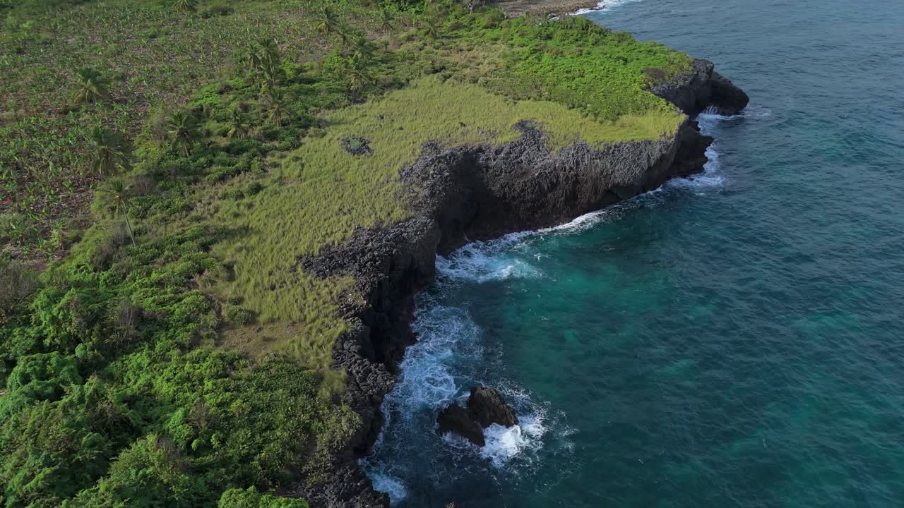 Aerial view of rugged coastal cliffs with turquoise waves and lush greenery in Las Galeras, Samana, Dominican Republic. Pan right
