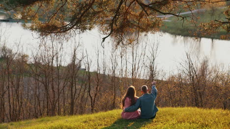 romantic couple enjoys the nature. young man and woman sitting on grass on the hill and looking at beautiful river in springtime. back view.