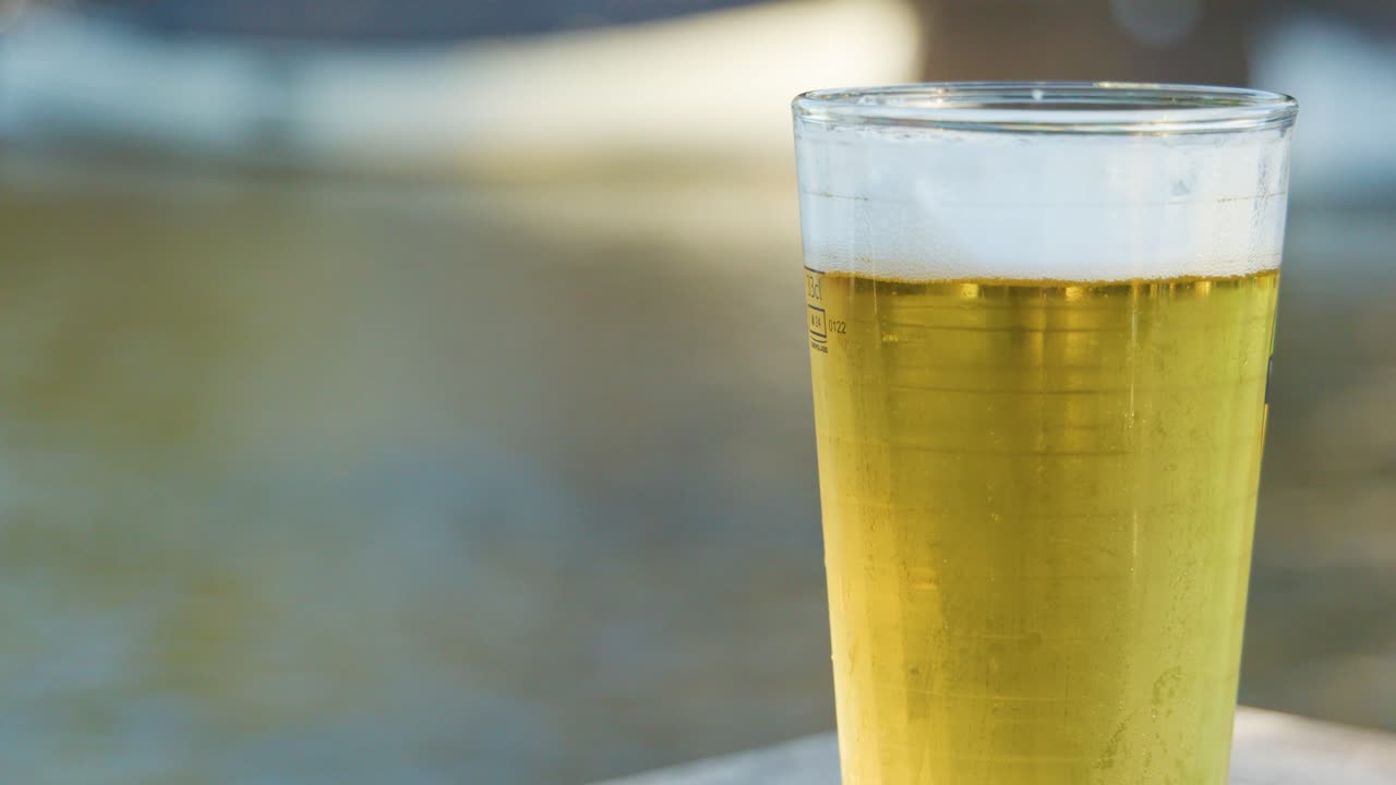 A hand reaches for and lifts a pint glass of pale beer from a wooden table outdoors, with a blurred river background and natural daylight