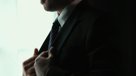 confident businessman in classic blue suit tying or adjust necktie near window in hotel room at the morning. handsome man wearing a nice suit on wedding day.