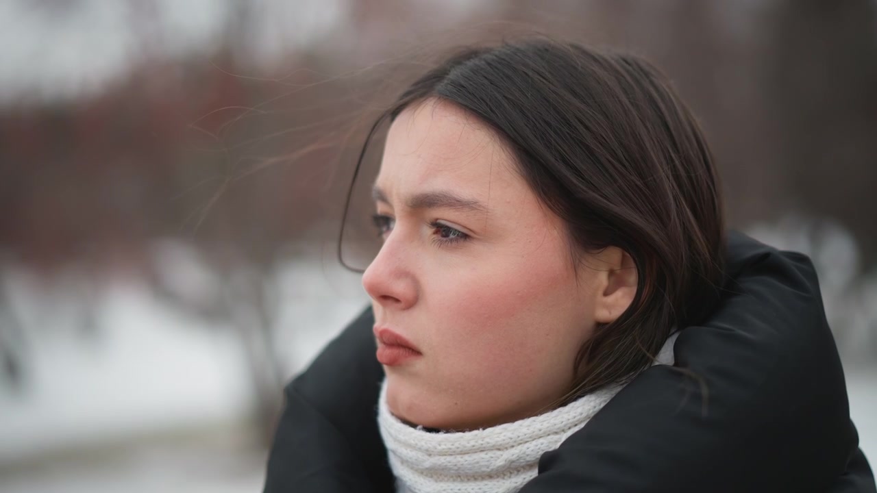 Close up of young woman wearing winter clothing outdoors in cold weather. Her hair is visible and woman appears to be looking around with slight head movement. background shows out of focus