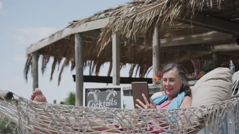 Happy-senior-caucasian-woman-lying-in-hammock-using-tablet-by-beach-bar,-in-slow-motion