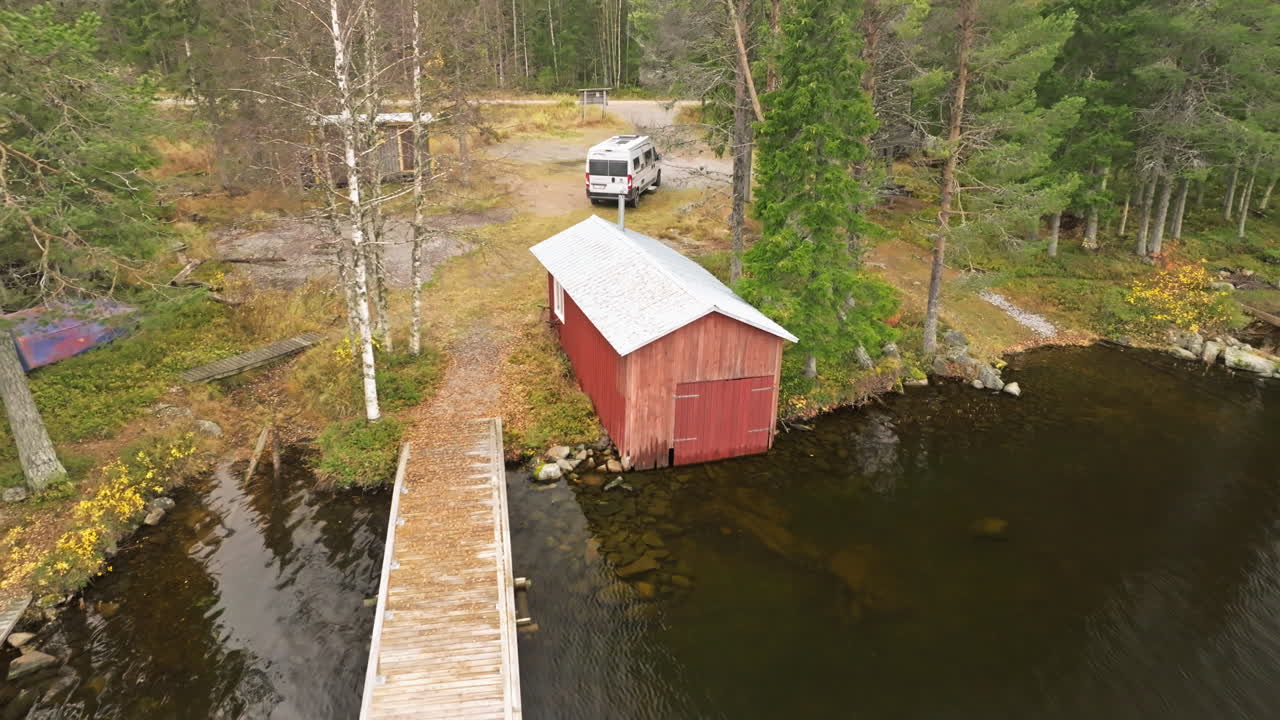 Svezia - Un sereno lago autunnale circondato da abeti e betulle con un camper bianco parcheggiato nelle vicinanze - Ripresa aerea in allontanamento