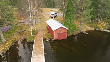 Suecia---Un-Sereno-Lago-Otoñal-Rodeado-De-Abetos-Y-Abedules-Con-Una-Autocaravana-Blanca-Estacionada-Cerca---Toma-Aérea-De-Retroceso